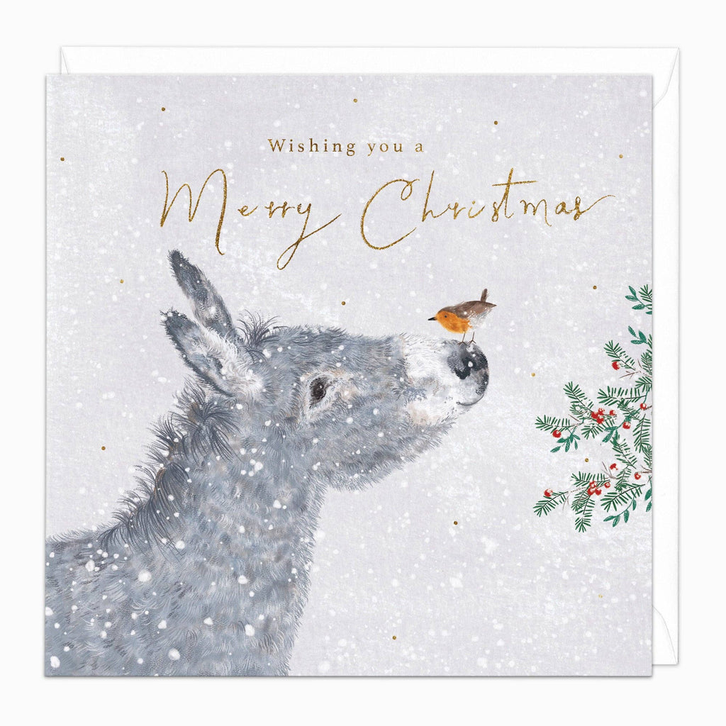 A square Christmas greeting card with a white speckled background, featuring a gray donkey, a small robin bird, green holly leaves, red berries, and gold script text reading 'Wishing you a Merry Christmas.' An envelope is partially visible behind the card.