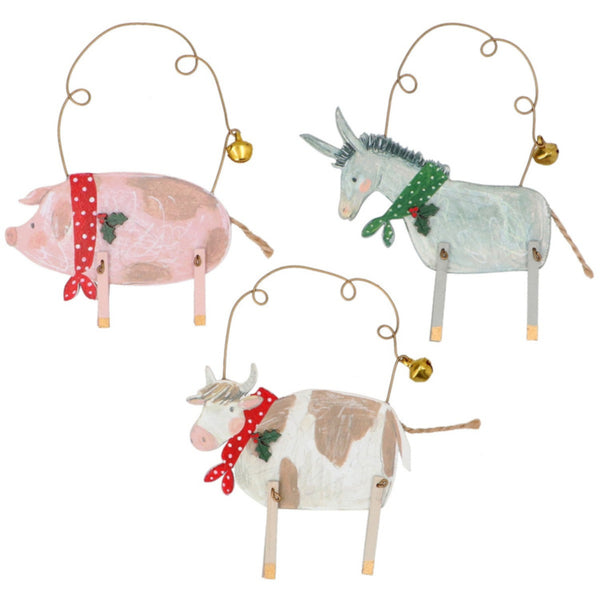 Three rustic Christmas animal ornaments hanging from wire loops: a pink-and-white pig with a red polka-dot scarf, a pale blue-gray donkey with a green scarf, and a white-and-tan cow with a red scarf, each with a small gold bell.