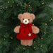 A plush teddy bear Christmas ornament with light brown fur wearing a red knit dress and a silver star, hanging on a green Christmas tree with warm string lights.