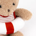 A close-up of a plush teddy bear with tan fur wearing a red and white striped shirt, holding a white life preserver with a red stripe, against a plain white background.