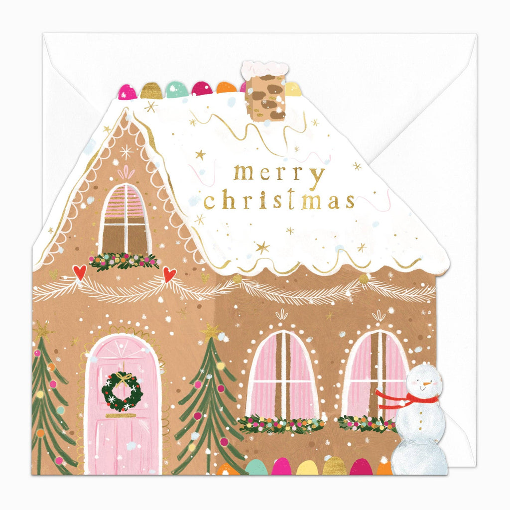 A Christmas greeting card featuring a gingerbread house with pink windows, a green wreath, snow-covered roof, gold 'merry christmas' text, a snowman, and colorful eggs, shown with a white envelope behind it.