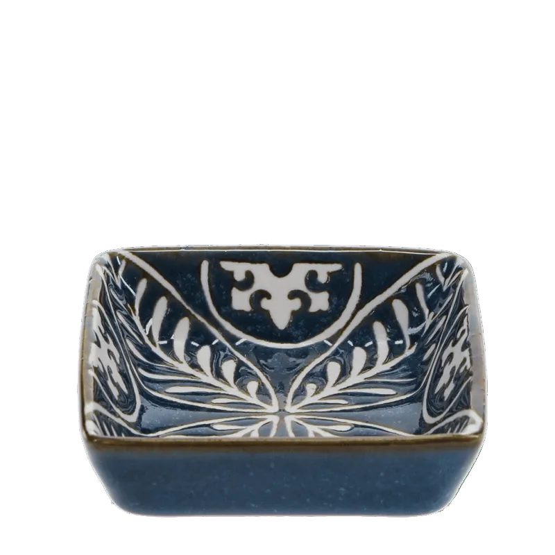 A square ceramic bowl with rounded corners, featuring a dark blue glaze and hand-painted white floral and scrollwork patterns, set against a solid green background.
