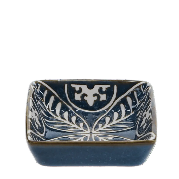 A square ceramic bowl with rounded corners, featuring a dark blue glaze and hand-painted white floral and scrollwork patterns, set against a solid green background.