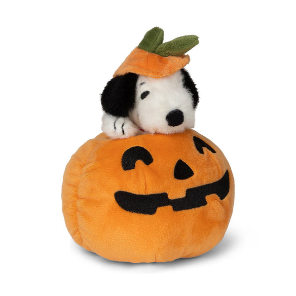 A plush toy featuring the Peanuts character Snoopy wearing an orange pumpkin hat with green leaves, sitting on top of a round orange jack-o'-lantern pillow with a black stitched face, photographed on a clean white background.