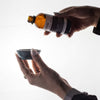 Hand holding a small container with another hand pouring a liquid from a bottle labeled 'Mystical Oil' on a white background.