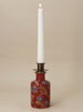 A tall white taper candle in a decorative red ceramic holder with floral detailing, set against a plain beige background.