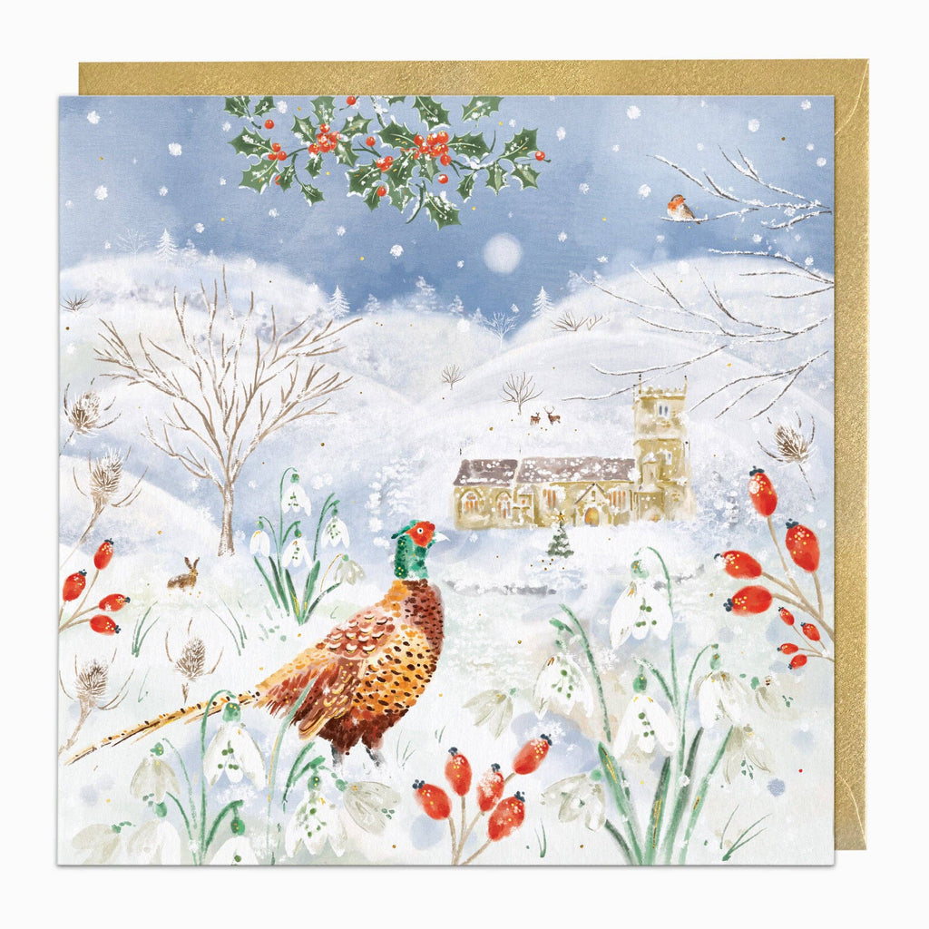 A square greeting card featuring a winter village scene with snow, bare trees, a church, red berries, and a pheasant in the foreground, shown with a gold envelope behind it.