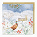 A square greeting card featuring a winter village scene with snow, bare trees, a church, red berries, and a pheasant in the foreground, shown with a gold envelope behind it.