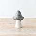 A small decorative mushroom figurine with a speckled dark gray cap and a smooth off-white stem, standing on a light wood surface against a plain white background.