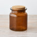 An amber glass jar with a cork stopper, set on a light wood surface against a plain white background.