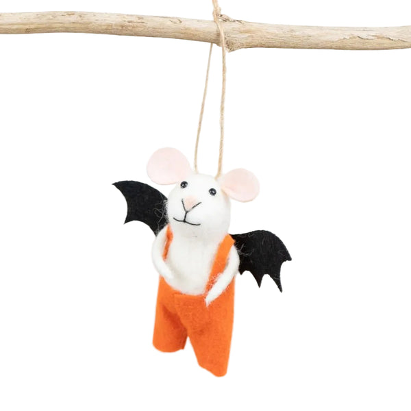 A small felt mouse hanging from a twine loop attached to a wooden branch, dressed in orange overalls with black bat wings, against a soft white and light wood background.