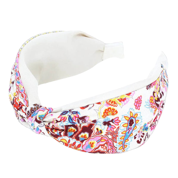 A wide fabric headband with a colorful paisley and floral print on a white base, featuring a solid white inner lining and a white adjustable tail tab, shown on a light gray background.