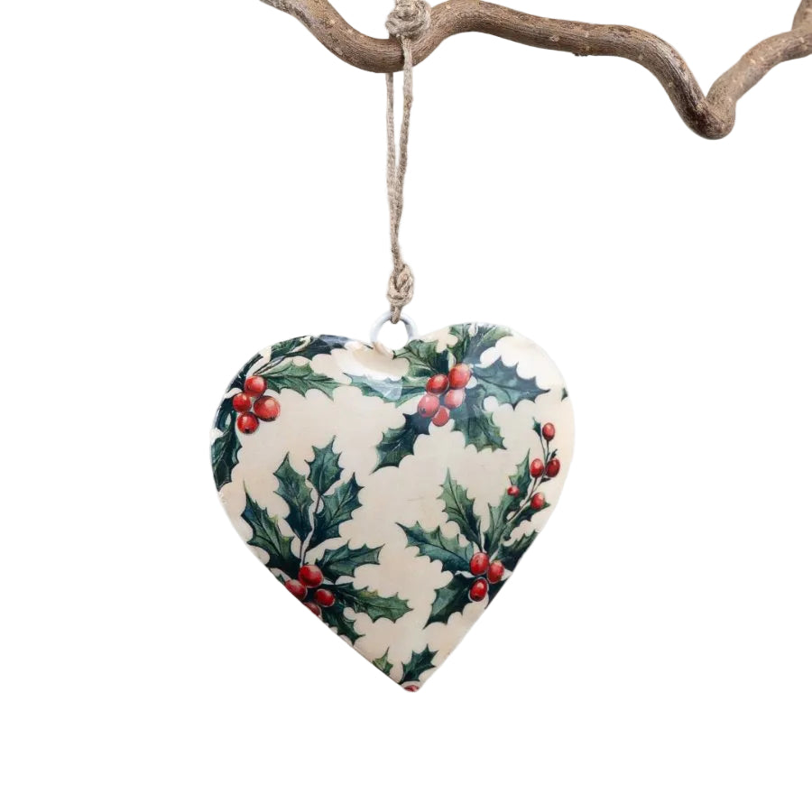 A heart-shaped Christmas ornament with a holly and berry pattern, hanging from a rustic branch against a clean white background.
