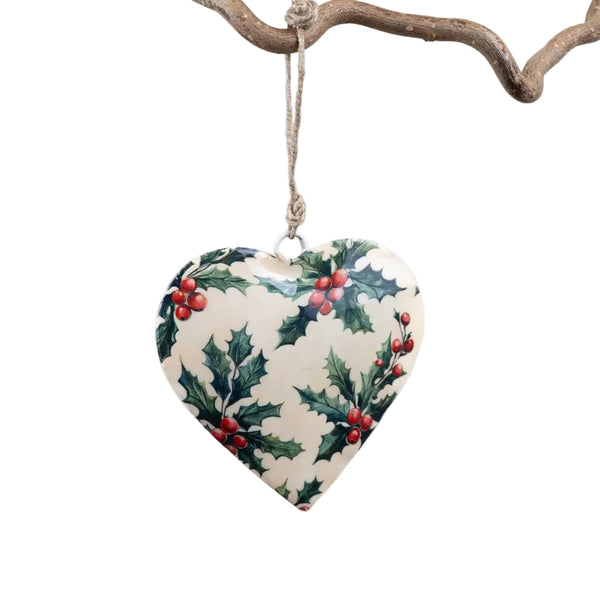 A heart-shaped Christmas ornament with a holly and berry pattern, hanging from a rustic branch against a clean white background.