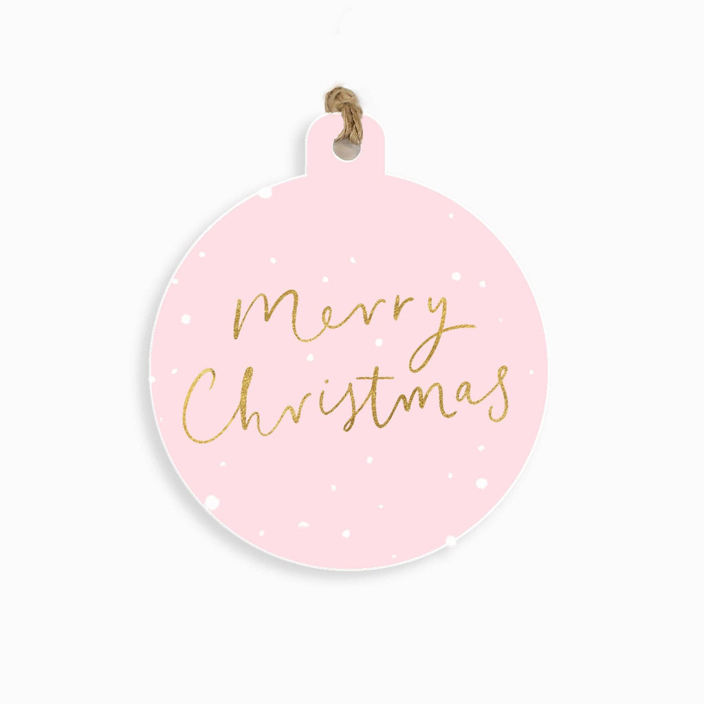 A round light pink Christmas ornament with 'Merry Christmas' written in gold cursive script, decorated with small white dots, and hanging from a natural jute twine loop.