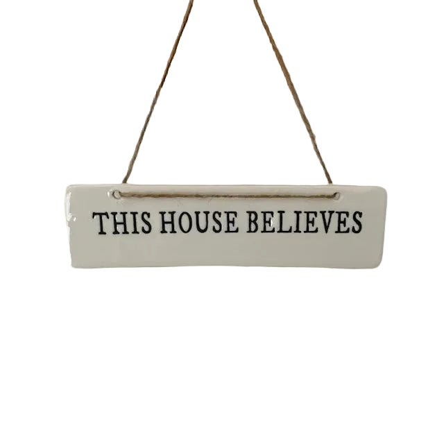 A white rectangular hanging sign with the phrase 'THIS HOUSE BELIEVES' in black lettering, suspended by twine from a wooden trim against a white background