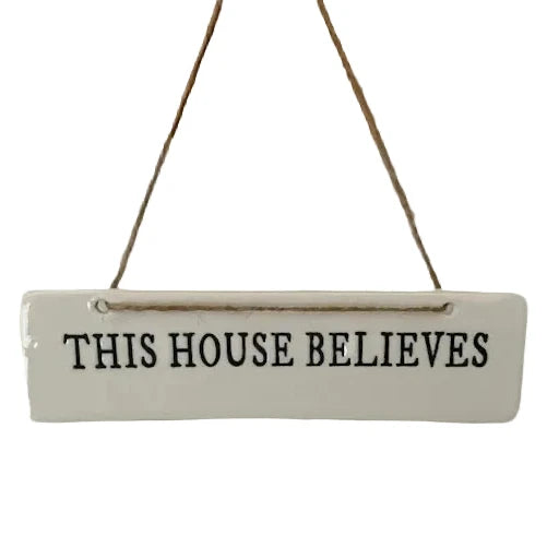A white rectangular hanging sign with the phrase 'THIS HOUSE BELIEVES' in black lettering, suspended by twine from a wooden trim against a white background