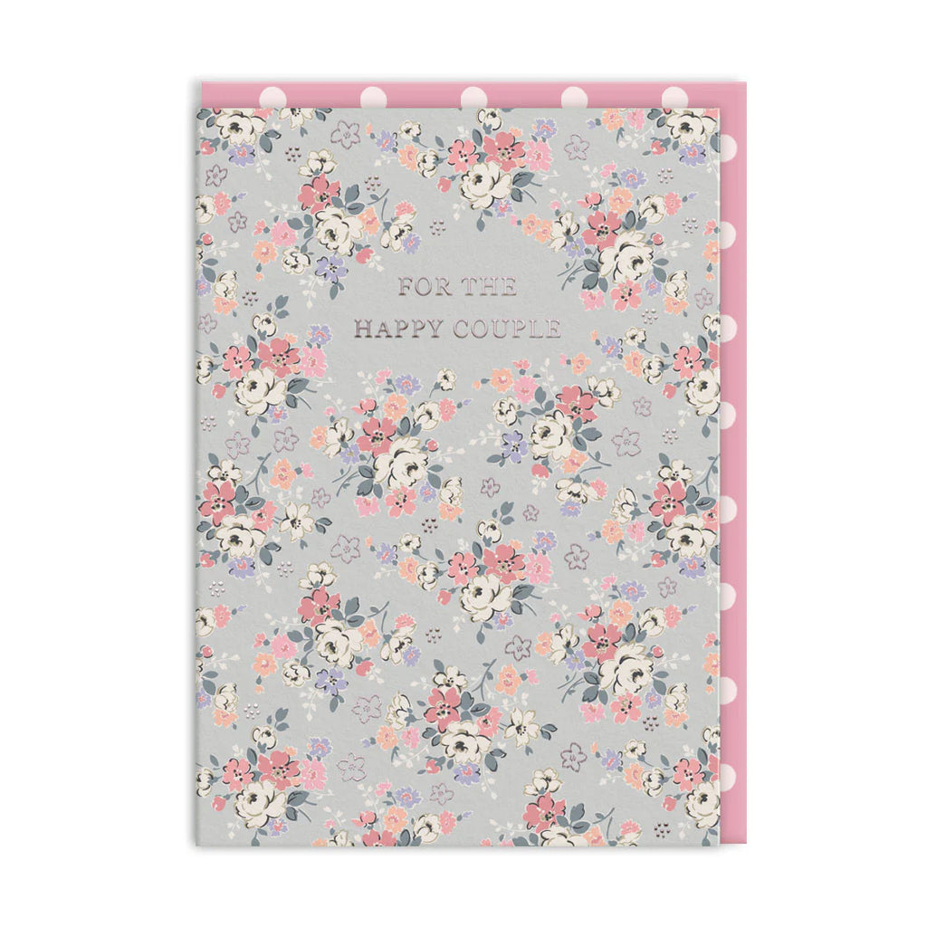 A floral greeting card with the text 'FOR THE HAPPY COUPLE' in the center, featuring a light gray background with pink, peach, white, and purple flowers. The card is shown with a pink backing board and white pom-pom trim along the top and right edges.