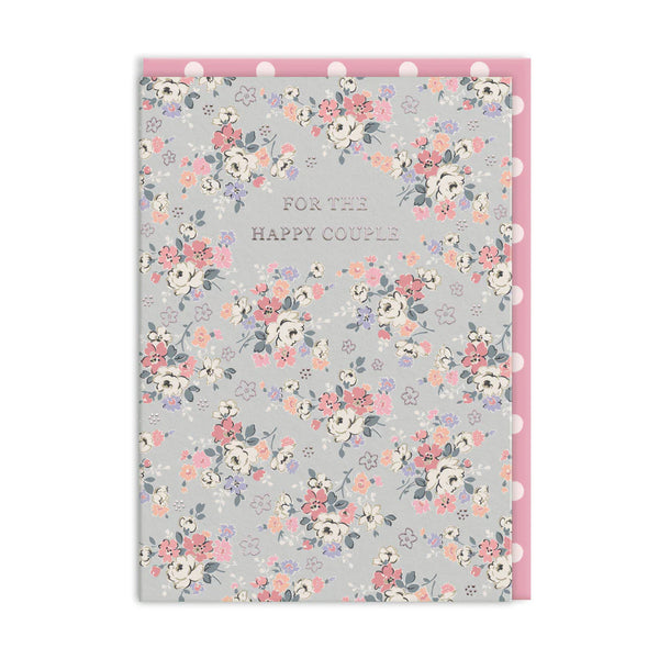 A floral greeting card with the text 'FOR THE HAPPY COUPLE' in the center, featuring a light gray background with pink, peach, white, and purple flowers. The card is shown with a pink backing board and white pom-pom trim along the top and right edges.