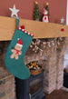 A teal felt Christmas stocking with a gingerbread man decoration hangs from a wooden mantel in a cozy fireplace setting. The mantel is decorated with white star cutouts, a small Christmas tree, a snowman figure, and a 'MERRY CHRISTMAS' banner.