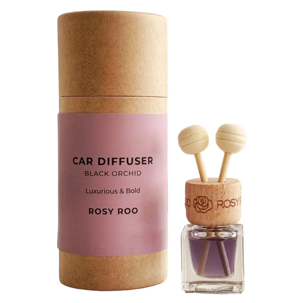 A car diffuser bottle with two wooden scent sticks and a clear glass base stands beside its kraft paper cylindrical packaging. The label is dusty pink with black text reading 'CAR DIFFUSER BLACK ORCHID Luxurious & Bold ROSY ROO'.