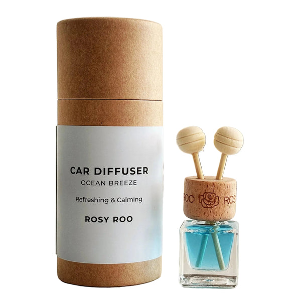 A car diffuser bottle with blue liquid and two wooden dropper tops stands beside its kraft paper cylindrical packaging on a light surface. The label reads 'CAR DIFFUSER OCEAN BREEZE Refreshing & Calming ROSY ROO'.