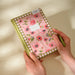 Floral notebook with a checkered border held by hands on a light background