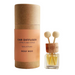 A car diffuser set on a light surface, featuring a tall kraft paper cylinder with a peach label reading 'CAR DIFFUSER CITRUS AND SPICE Spicy & Exotic ROSY ROO' next to a small clear glass bottle with yellow liquid and two wooden diffuser sticks. A neutral vase with dried stems sits blurred in the background.