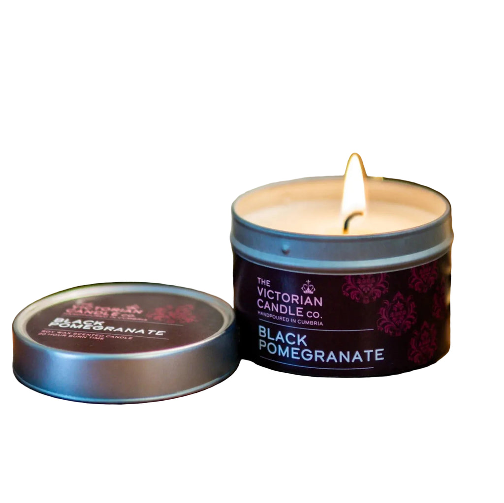 Candle in a tin labeled 'Black Pomegranate' from The Victorian Candle Co. on a wooden surface with a blurred background.