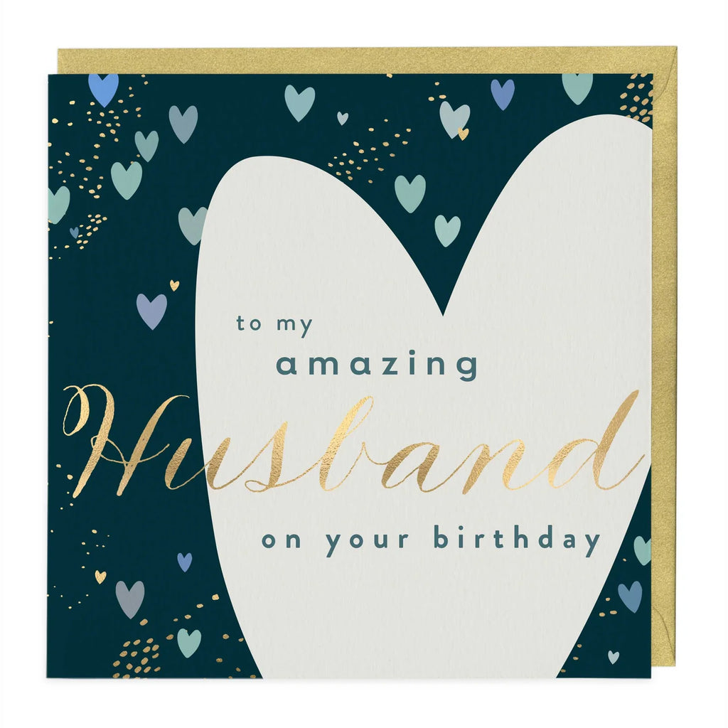 A square birthday card with a gold envelope behind it. The card has a dark teal background decorated with small pastel hearts and gold confetti, featuring a large white heart shape and the text 'to my amazing Husband on your birthday' in teal and gold lettering.