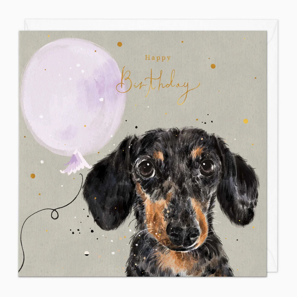A square birthday card featuring a watercolor-style illustration of a black and tan dachshund with a light purple balloon and gold script reading 'Happy Birthday' on a warm gray background.