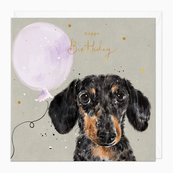 A square birthday card featuring a watercolor-style illustration of a black and tan dachshund with a light purple balloon and gold script reading 'Happy Birthday' on a warm gray background.