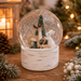 A festive Christmas snow globe with a birch-wood-textured base, featuring a small green tree, a beige deer, and a white rabbit inside glittery white snow, displayed on a wooden surface with warm holiday lights in the background.