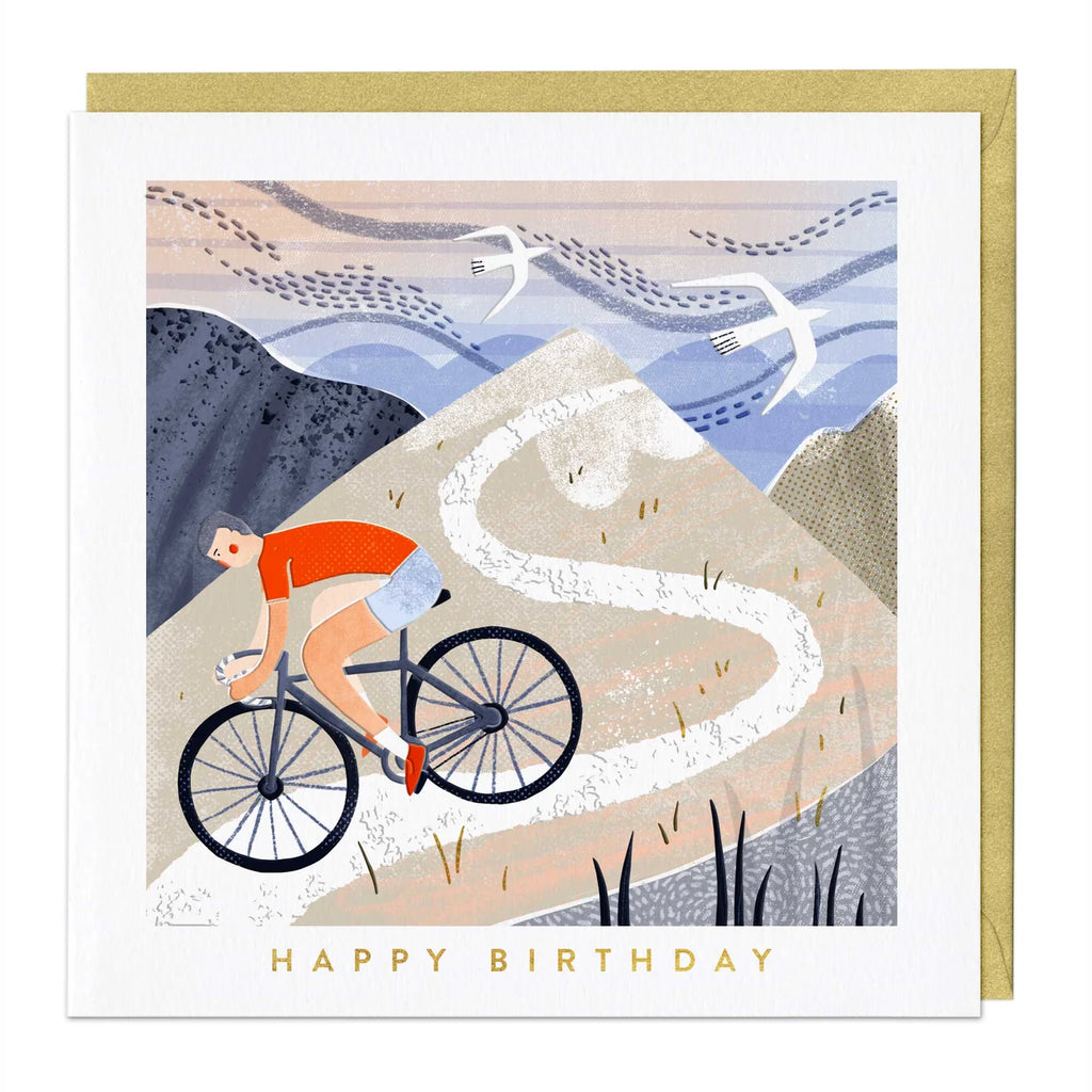 A square birthday card with a gold envelope behind it, featuring an illustrated cyclist riding up a winding mountain path. The card reads 'HAPPY BIRTHDAY' in gold text at the bottom.
