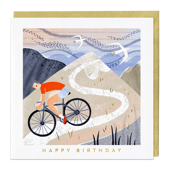 A square birthday card with a gold envelope behind it, featuring an illustrated cyclist riding up a winding mountain path. The card reads 'HAPPY BIRTHDAY' in gold text at the bottom.