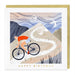 A square birthday card with a gold envelope behind it, featuring an illustrated cyclist riding up a winding mountain path. The card reads 'HAPPY BIRTHDAY' in gold text at the bottom.