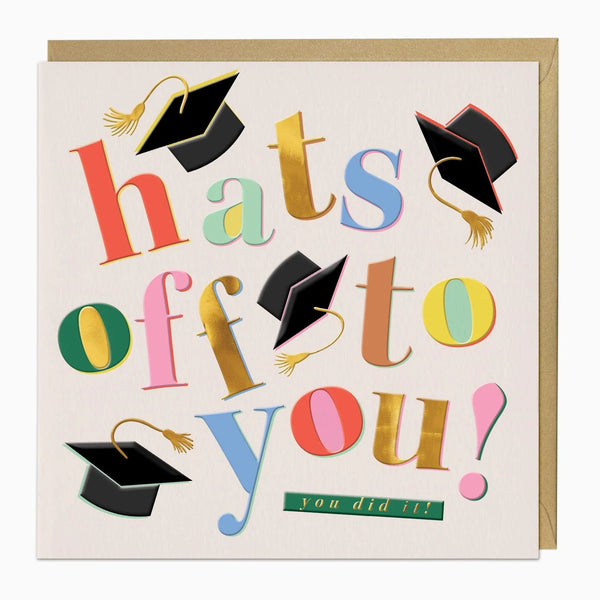 A square graduation greeting card with a kraft envelope behind it. The card reads 'hats off to you!' in large multicolored playful letters, with several black graduation caps illustrated around the words and a small green banner that says 'you did it!'.