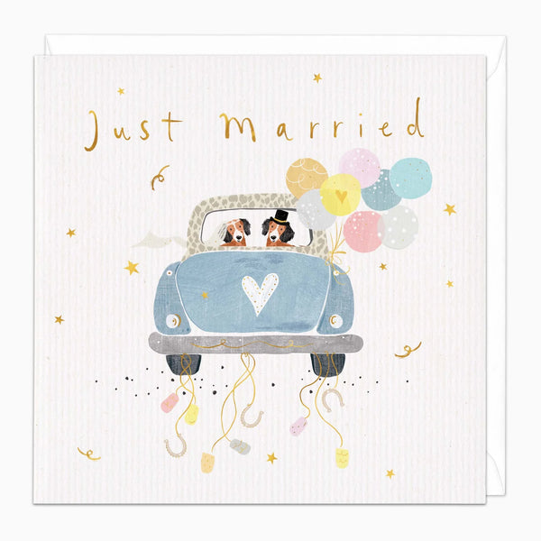 Square greeting card with a white textured background featuring the words 'Just Married' in gold script. The center illustration shows a light blue car with a white heart on the front, driven by two dogs wearing top hats, with pastel balloons and confetti falling below.