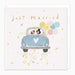 Square greeting card with a white textured background featuring the words 'Just Married' in gold script. The center illustration shows a light blue car with a white heart on the front, driven by two dogs wearing top hats, with pastel balloons and confetti falling below.