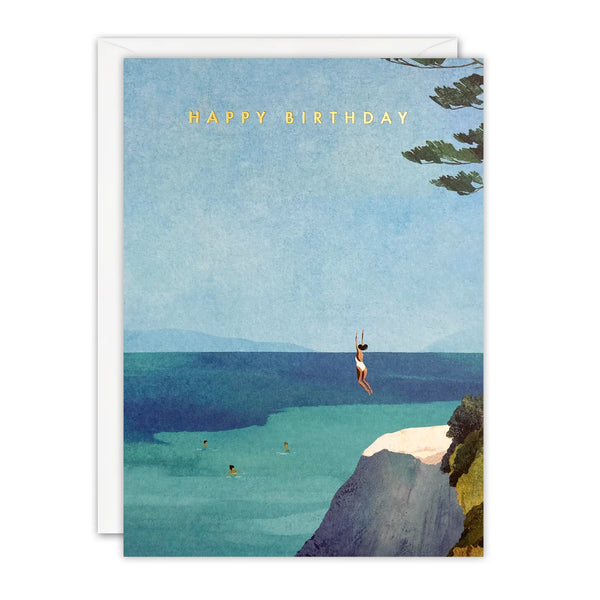 A square birthday card with a white envelope behind it, featuring an illustrated beach scene with a person hanging from a swing over the ocean and the text 'HAPPY BIRTHDAY' in gold at the top.