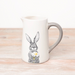 A ceramic serving jug featuring a hand-drawn illustration of a hare (or rabbit) holding a daisy on a textured background.