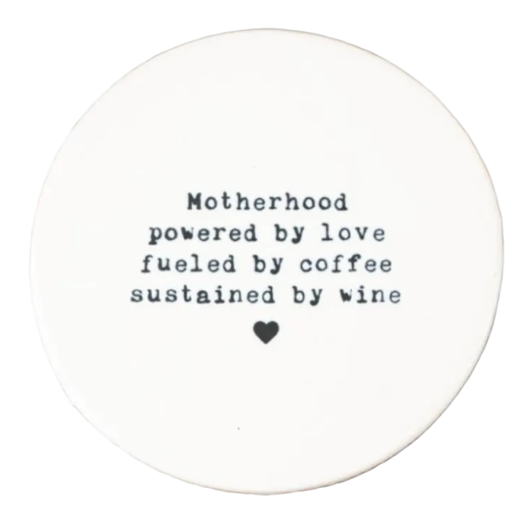 A porcelain coaster featuring a hand-stamped, typewriter-style font with the quote: "Motherhood powered by love fueled by coffee sustained by wine," finished with a small black heart at the bottom on a white background.