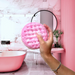Hand holding a pink exfoliating pad in a bathroom with pink marble walls and a pink bathtub.