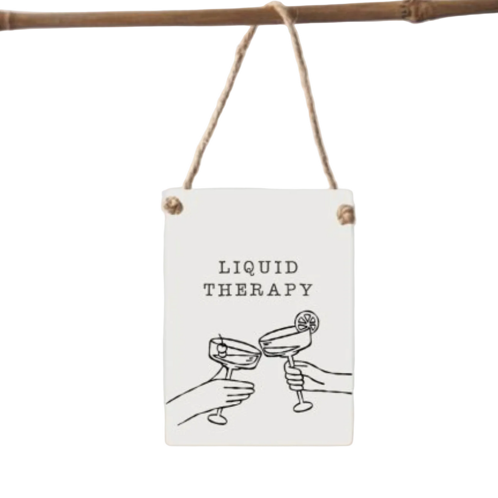 A rectangular white hanging sign with a rustic twine loop and wooden grommets, featuring the text 'LIQUID THERAPY' above a simple black line drawing of two hands clinking cocktail glasses. The sign hangs from a natural wood rod against a light gray background.