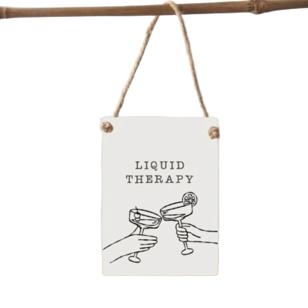 A rectangular white hanging sign with a rustic twine loop and wooden grommets, featuring the text 'LIQUID THERAPY' above a simple black line drawing of two hands clinking cocktail glasses. The sign hangs from a natural wood rod against a light gray background.