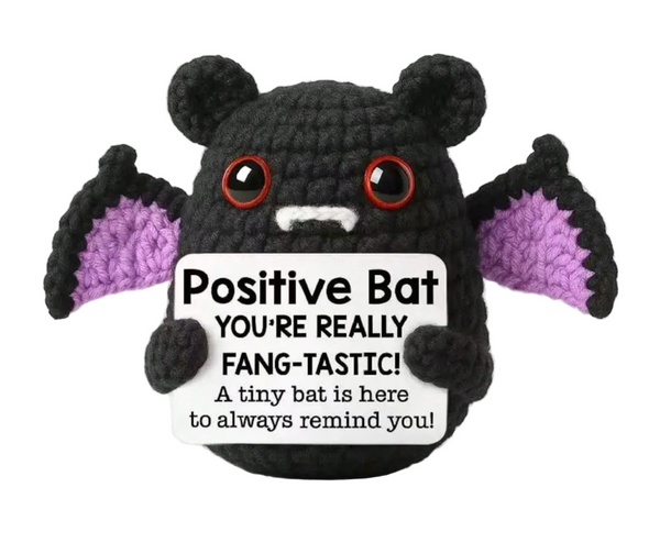 A crocheted black bat plush toy with purple wings holds a white sign that reads, 'Positive Bat YOU'RE REALLY FANG-TASTIC! A tiny bat is here to always remind you!' on a plain white background.