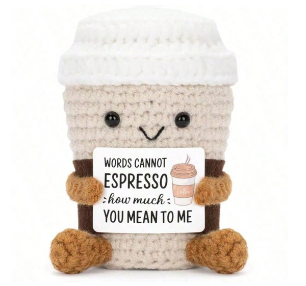 A crocheted coffee cup character plush toy holding a sign that reads 'WORDS CANNOT ESPRESSO how much YOU MEAN TO ME' with a small coffee cup illustration. The cup is off-white with brown yarn arms and feet, and black embroidered facial features, shown on a plain white background.