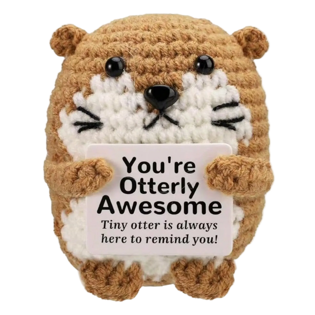 A crocheted otter plush toy holding a white sign that reads 'You're Otterly Awesome Tiny otter is always here to remind you!' on a plain white background.