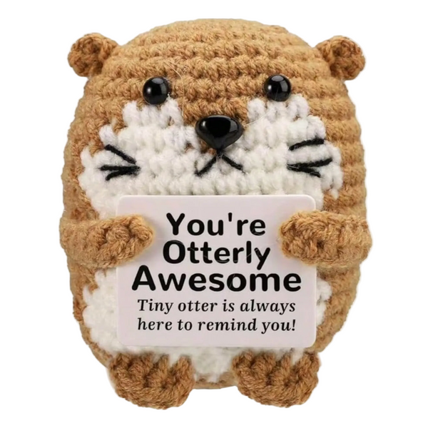 A crocheted otter plush toy holding a white sign that reads 'You're Otterly Awesome Tiny otter is always here to remind you!' on a plain white background.