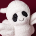 A close-up of a white plush ghost toy with black embroidered eyes and a smiling mouth, set against a solid dark red background.